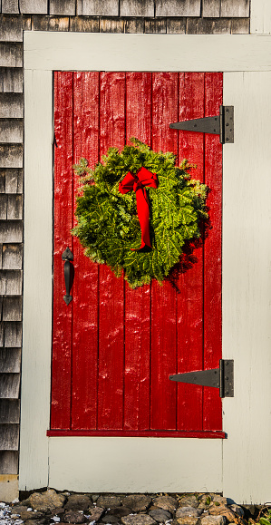 A balsam Christmas wreath with a red ribbon hangs on a red barn door on Cape Cod.