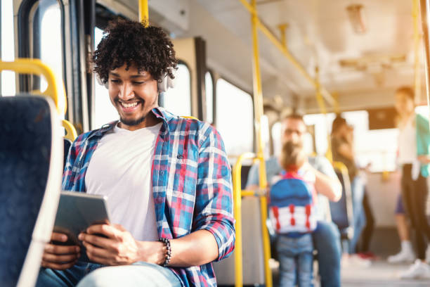 lächelnde afrikanische amerikanische mann sitzen in öffentlichen verkehrsmitteln und mit tablet. auf ohren kopfhörern. im hintergrund die leute sitzen und stehen. - bussing stock-fotos und bilder