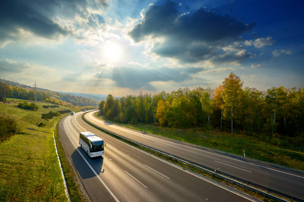 istock Three white buses traveling on the asphalt highway between deciduous forest in autumn colors under the radiant sun and dramatic clouds. View from above. 1082146588