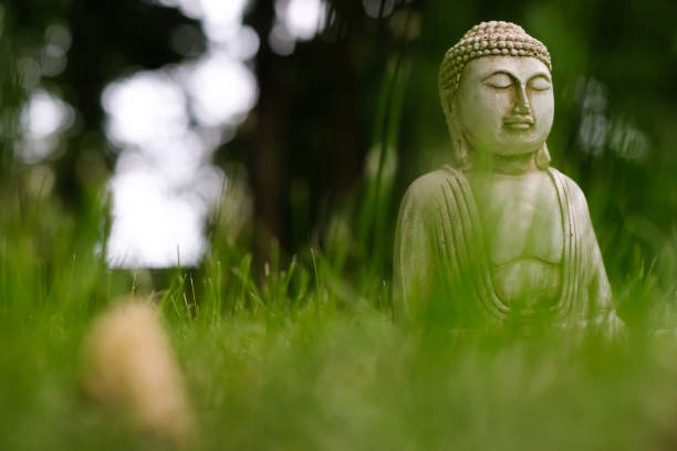 Small white Buddha statue in a meditation pose with green grass foreground and in natural bright blurred background. Religious symbol of buddhism. Selective focus.