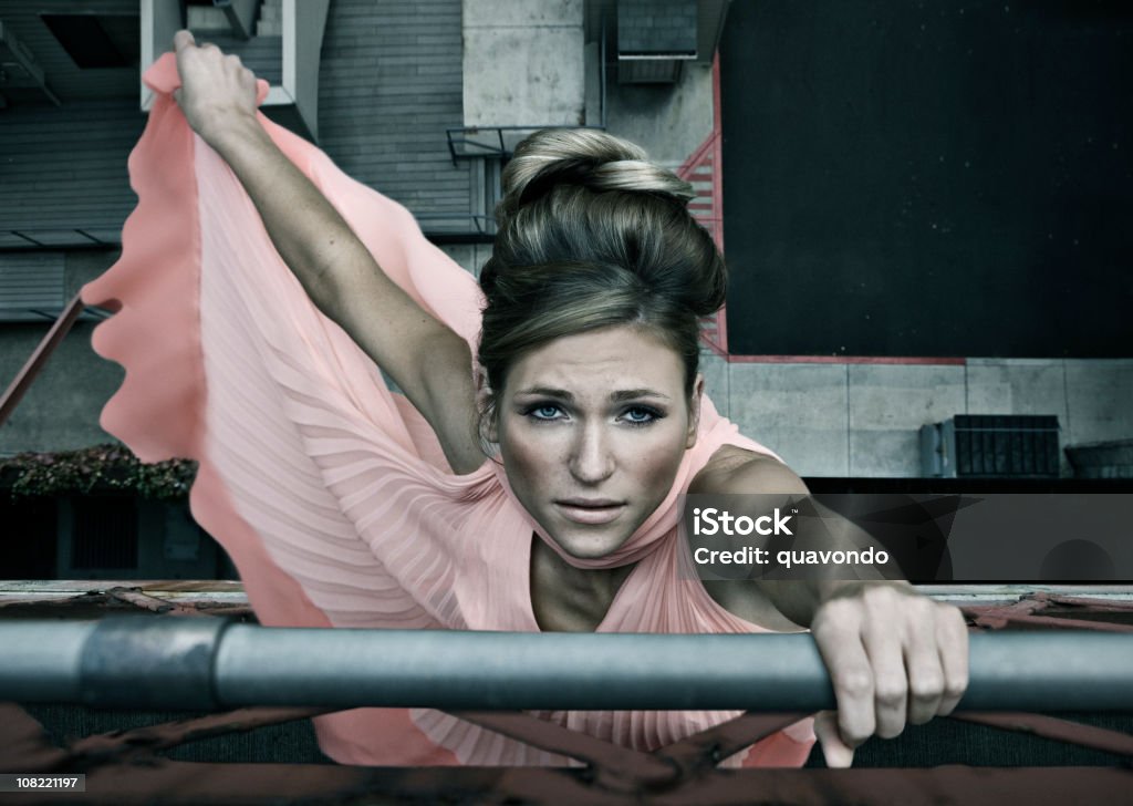 Beautiful Young Woman Fashion Model in Dress Hanging Off Bridge Caucasian fashion model with pink sleeveless dress hanging onto building. Falling Stock Photo