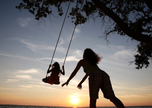 girl of five on swing. She sits pensively and looks up. Green trees in background.
