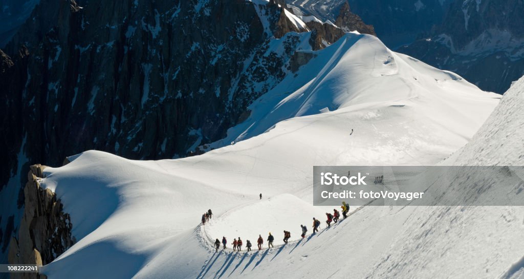 Group of People Climbing Snow Covered Mountain Intrepid Alpinists carefully negotiating the steep snow ridge from the Aiguille du Midi high on the Mont Blanc massif high in the to the glacial col of the Vall Climbing Stock Photo