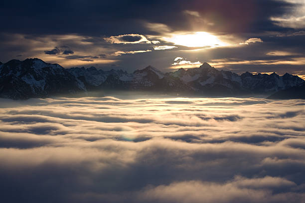 A dramatic landscape where a dark silhouette of a mountain intersects with intense backlighting. This energetic landscape photo emphasizes lens flare and light rays. A dynamic depiction of natural scenery with the sun peeking through gaps in the clouds.