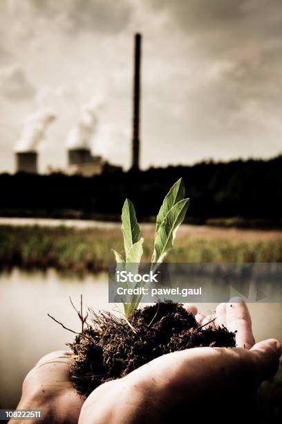 Person Holding Plant Seedling With Factory In Background Stock Photo - Download Image Now