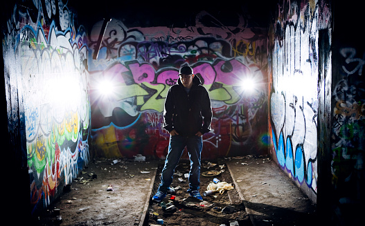 A young man in hip hop rap fashion stands in front of a graffiti covered city wall at night.