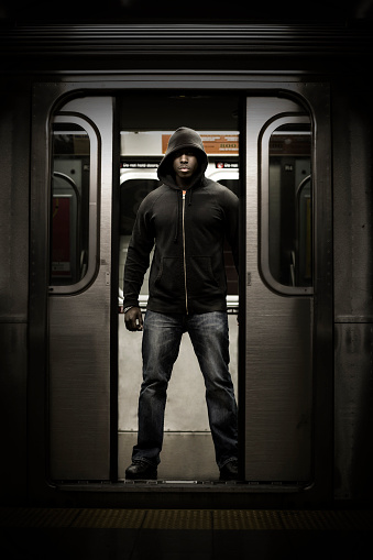 Intimidating Black male wearing hooded sweatshirt standing on subway train as doors open.