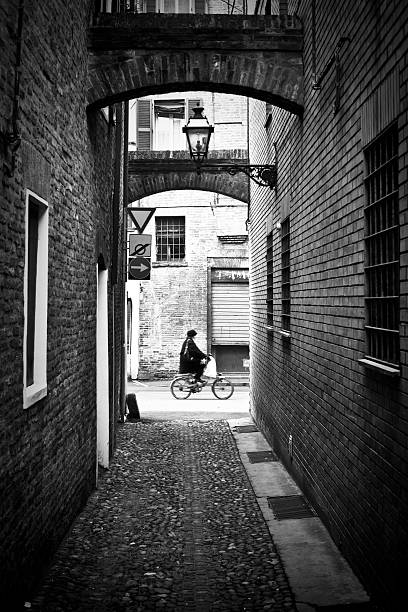 An old woman is cycling at the end of a dark and narrow alley. Soft motion. This mediveal area of the city is an Unesco World Heritage Site. Location: Ferrara, Italy.