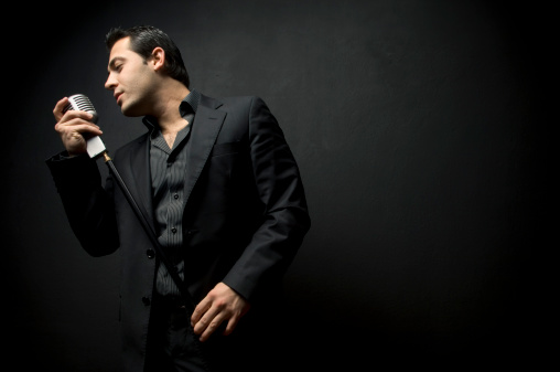 Black suit weared male singer singing with old style microphone in front of a black wall.