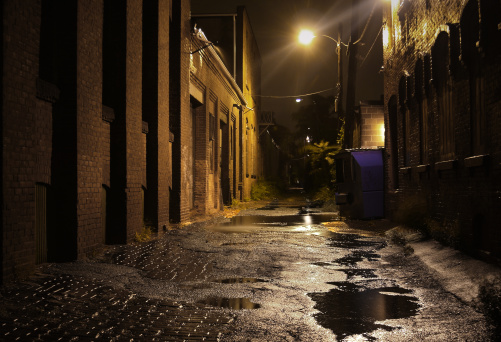 A deserted alley in a park with a stone surface and metal benches along the edge on a rainy night. The light from the street lamps reflects off the wet stone surface.