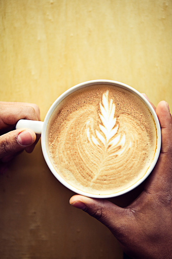 A pair of hands cradling a warm mug of foamy latte, complete with froth art.  Vertical with copy space.