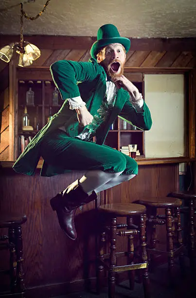 A "Leprechaun" looking man with red hair and beard, in a full green suit complete with vest, bow tie, and top hat, perfect for St. Patrick's Day.  He dances and jumps in the air at an old fashioned Irish bar. Vertical with copy space.