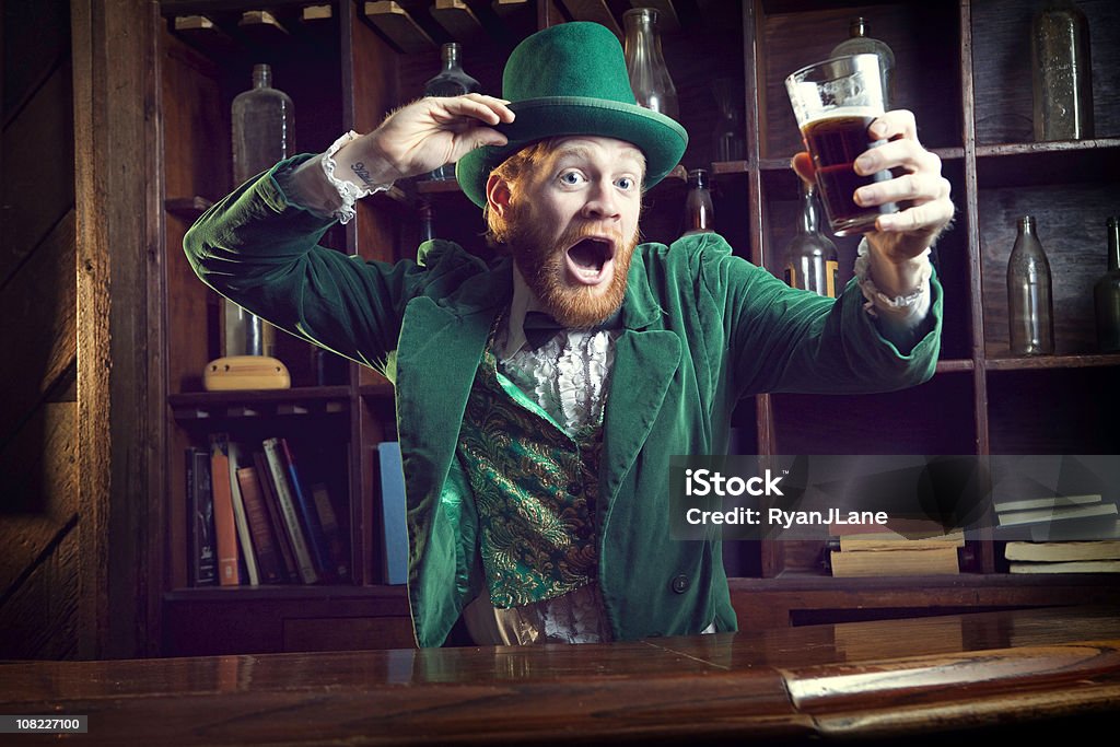 Irish Character / Leprechaun Celebrating with Pint of Beer A "Leprechaun" looking man with red hair and beard, in a full green suit complete with vest, bow tie, and top hat, perfect for St. Patrick's Day.  He stands at an old fashioned Irish bar with a glass of beer in his hand dancing and tipping his hat.  Horizontal with copy space. St. Patrick's Day Stock Photo