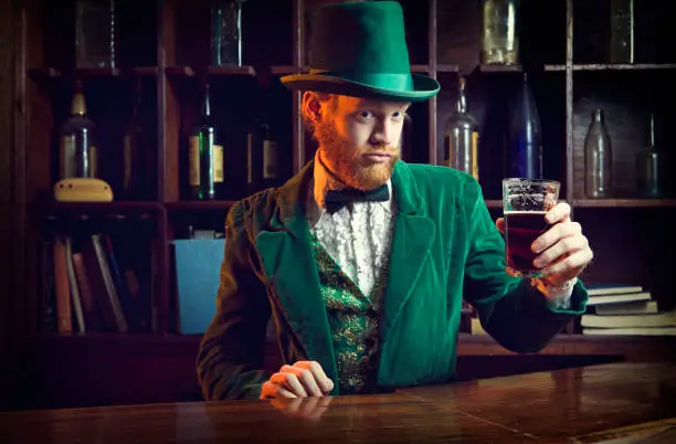 A "Leprechaun" looking man with red hair and beard, in a full green suit complete with vest, bow tie, and top hat, perfect for St. Patrick's Day.  He stands at an old fashioned Irish bar with a glass of beer in his hand.  Horizontal with copy space.