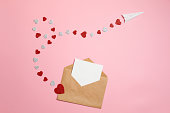 Distance love concept, sending love letter, valentines day. Kraft envelope with blank postcard and paper airplane flying on route made of heart shaped valentines cards lay on pink background desk