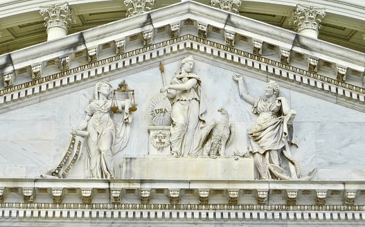 Close up America pediment sculpture (figures of America, Justice and Hope) above east central front of U.S. Capitol Building in Washington, DC. The central figure represents America, who rests her right arm on a shield inscribed 