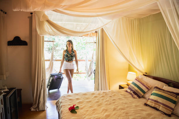 Woman in her 20s, pulling wheeled suitcase, entering luxury bedroom suite, with canopy over double bed