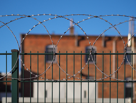 Green steel fence with barbed wire on top and old factory building with red brick walls in the background (out of focus)
