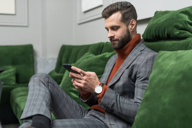 istock focused handsome man in formal wear sitting on green sofa and using smartphone 1082375700