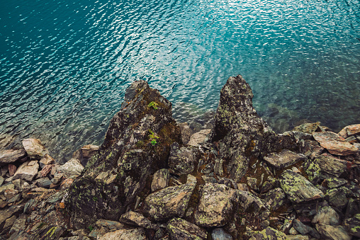 Rocky shore. Water edge. Shiny surface of azure mountain lake. Stony bottom in transparent water. Minimalist blue background. Reflection of mountains on clear water. Vegetation on stones. Copy space.