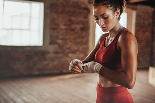Healthy young female boxer wearing strap on wrist. Muscular built woman getting ready for boxing exercise at fitness studio.