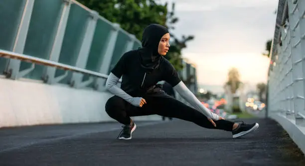 Hijab girl exercising on walkway bridge in early morning. Muslim woman wearing sports clothes doing stretching workout outdoors.