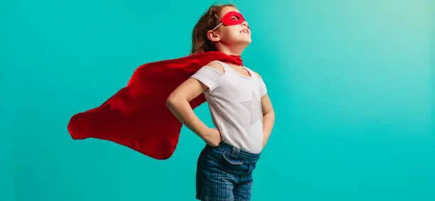 Girl child wearing red superhero costume standing with her hands on hips in studio. Girl in red cape and mask looking away on blue background.