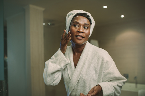 Reflection of african woman in bathroom mirror applying cosmetic cream on her face. Mature female putting on moisturizer on her face skin.