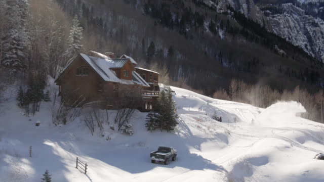 Aerial Drone Shot of a Person Snowblowing in Front of a Picturesque, Snow-Covered Mountain Cabin on a Bright Winter Day