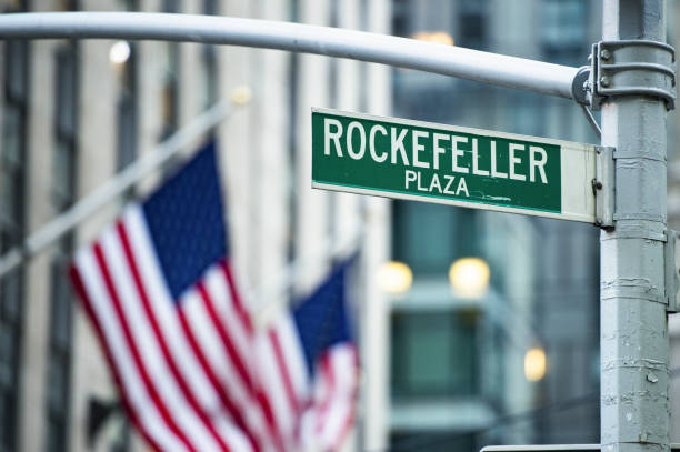 (selective focus) close-up view of green street sign depicting 'rockefeller plaza' in midtown manhattan, new york. blurred american flags in the background. - symbol finance corporate business manhattan imagens e fotografias de stock