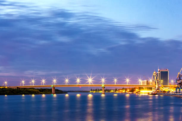 Road concrete bridge on supports across the Volga River, lampposts illuminate at dusk at night, green forest and industrial dock on different shores.