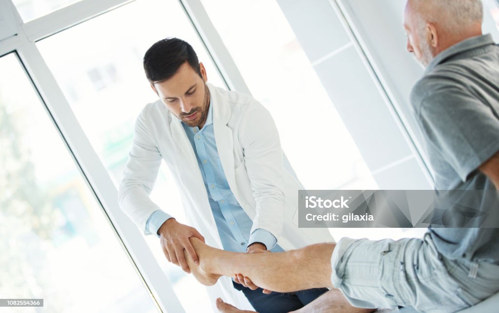Ankle examination. Closeup side view of an early 60's senior gentleman having some pain in his ankle. He's at doctor's office having medical examination by a male doctor. The doctor is gently twisting and moving the ankle to see what is the cause of pain. Doctor Stock Photo