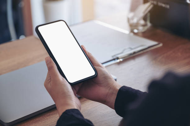 istock Mockup image of hands holding black mobile phone with blank white screen with laptop on wooden table 1082643554