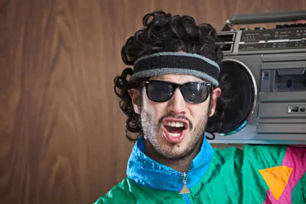 A hip funky young man with a mullet and fluorescent jump suit windbreaker.  Dark 80's style glasses cover his eyes as he sings along to his "ghetto blaster" boom box.  Wood paneled wall for copy space in the background.