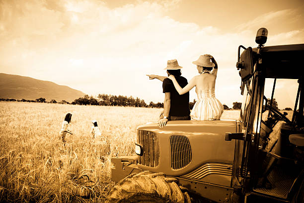 pareja sentada en tractor mientras los niños juegan - sepia toned field wheat sign fotografías e imágenes de stock