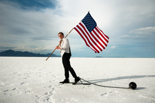 Young American businessman determined to move foward under difficult conditions symbolized by the desert and by the ball and chain.  Head up and looking at the camera and carrying an American flag.  Red Rockalypse 4 North.