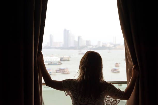 Asian woman opens window curtains with boats and bay background stock photo