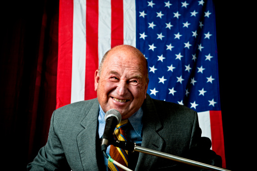 Portrait of an Adult Male in a Grey Suit, United States Elections Voter, Posing for Camera, Smiling. Caucasian Senior Man Standing in a Modern Polling Station with American Flag in the Background