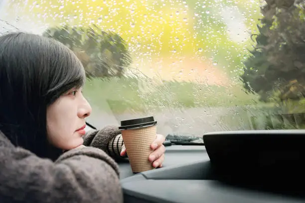 Rainy Day or Bad Weather in a Vacation Concept. a Sadness Woman with Hot Coffee sitting in Car and Looking Outside through Window, Waiting for Rain to stop