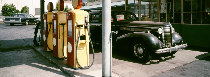 A gas station with 1940s fuel pumps and a vintage car. The photo is shot with a panoramic camera (Hasselbad XPAN), 45mm lens. The image is slightly toned to bring out the reds and yellows of the pumps.