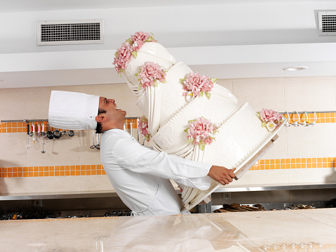 Young male pastry chef carrying a huge wedding cake in a commercial kitchen