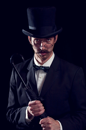 A sophisticated and classy looking man in a top hat and spectacles poses for a portrait.  Isolated on a black background.