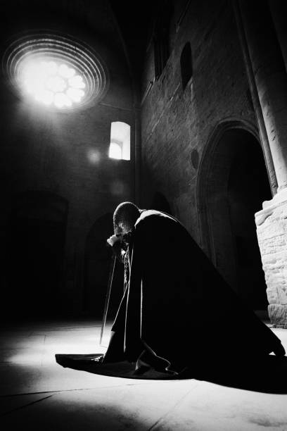 Dedication Knight Portrait Black & White High Contrast Strong Grain Dedication. Pensive knight kneeling in ancient cathedral holding his excalibur like sword. Natural light falling through the ancient window lighting up the scene. Medieval Knight Scene. High Contrast, BW, Intended Grain. church black and white people praying stock pictures, royalty-free photos & images