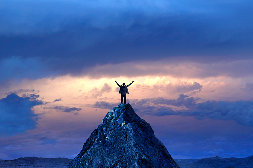 A silhouetted businessman stands on top of a mountain peak with outstretched arms in front of a dramatic sunset in the distance.