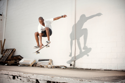 Afro-american young man riding skateboard making boardslide or lipslide stunt jumping up high outdoors in business area near glass building at sunny day. Extreme sports, active lifestyle, hobby.