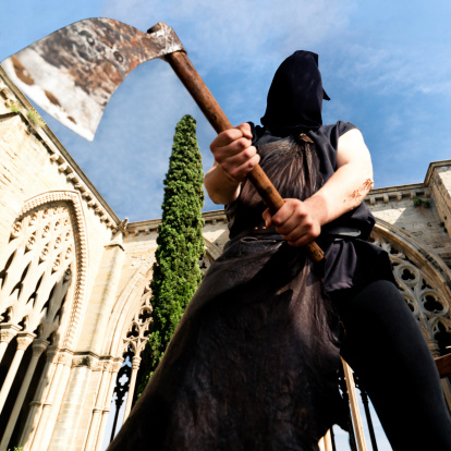 Headsman with axe standing in the patio of middle age monument. View from below against the blue sky. Selective focus on mask. Squared.