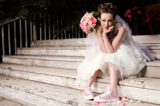 Beautiful Young Bride In Wedding Dress and Pink Sparkly Tennis Shoes.  Holding Pink and White Bouquet.  Smiling and Looking At Camera.  Sitting On Wooden Stairs. Dark Shadowy Background.