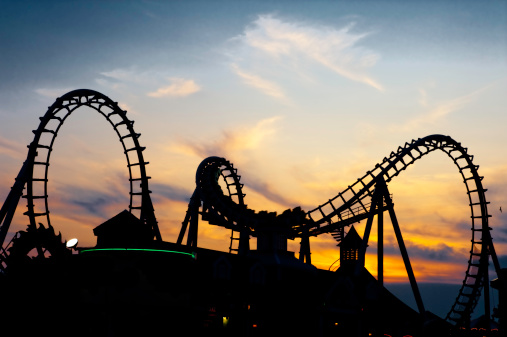 A black roller coaster is silhouetted against a vibrant orange and pink sunset sky in an amusement park