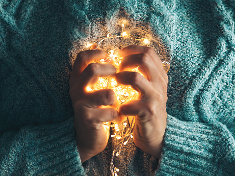 Warm heart, love concept. Women's hands holding a garland. Girl in a blue sweater with Christmas lights in her hands