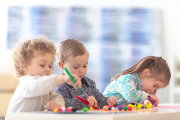 Three toddlers in preschool are sitting at a table and playing with arts-and-crafts supplies.